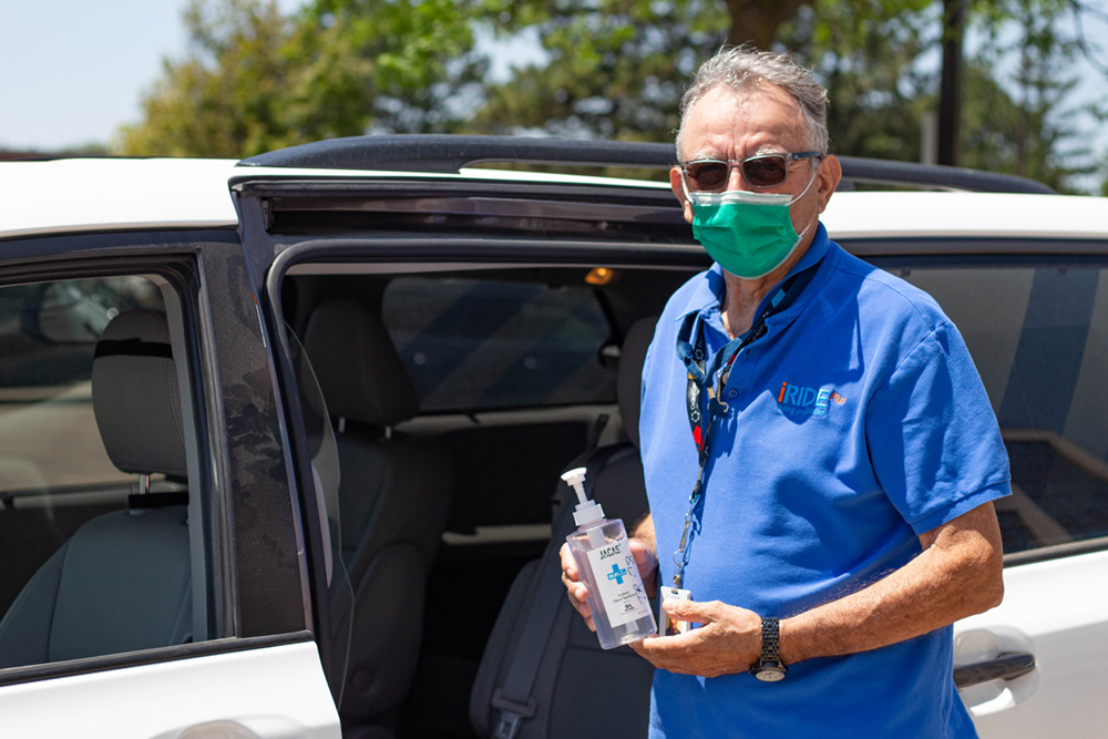 iRIDE driver wearing a mask holds up a bottle of hand sanitizer, and stands outside a van with the back door open.